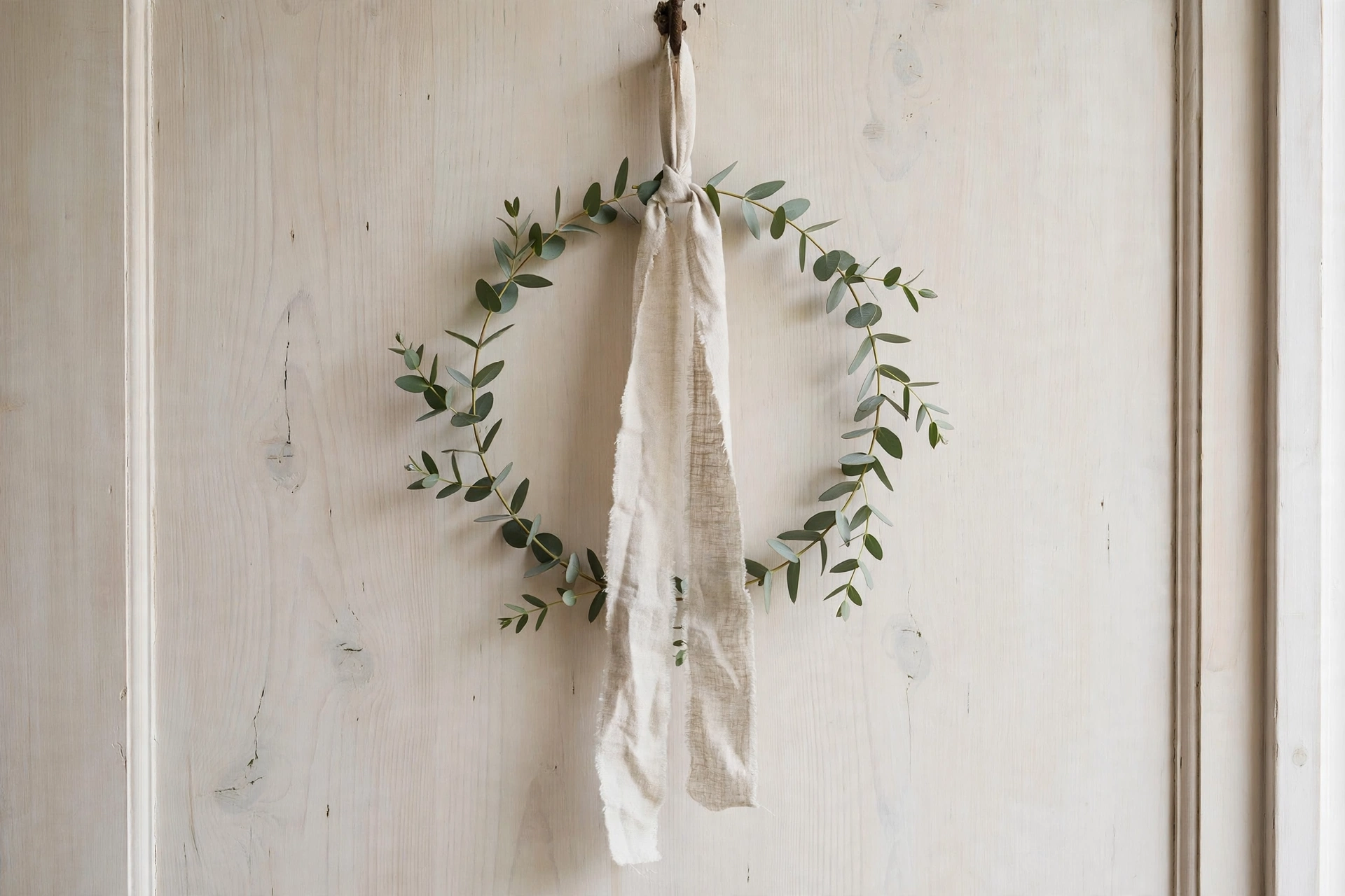Minimalist eucalyptus wreath with linen ribbon hanging on a pale wooden door.